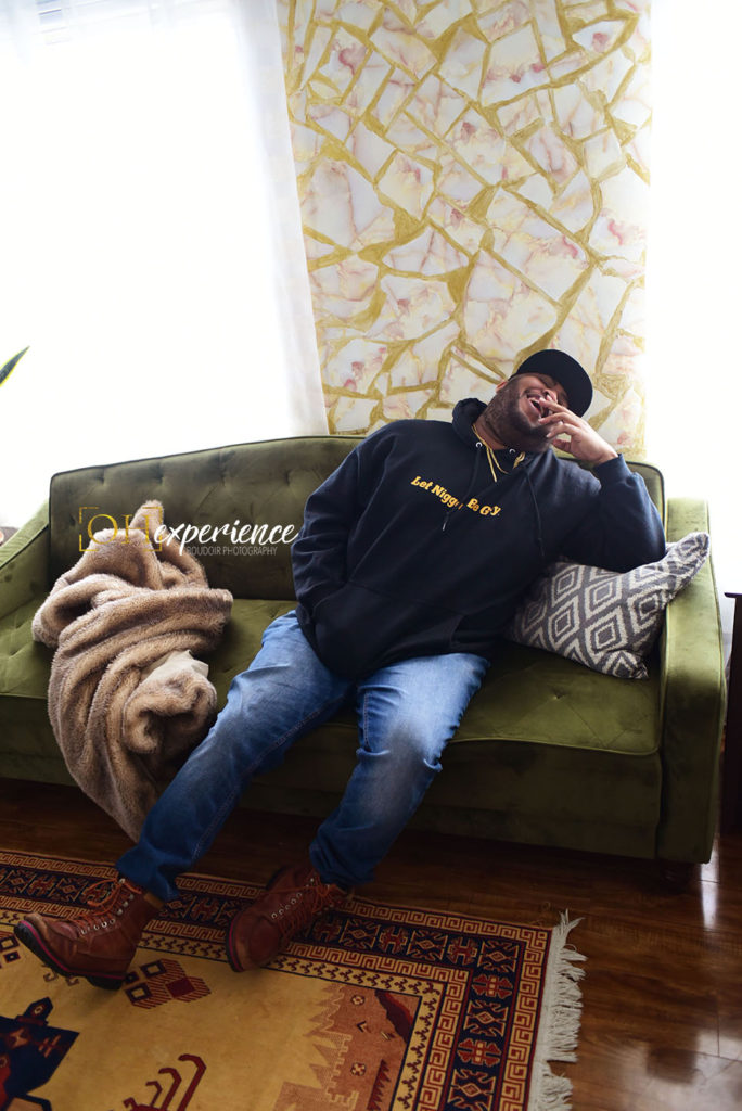 Black man sitting on a green sofa laughing. His hoodie reads, Let Niggas Be Gay. The Negus Project 