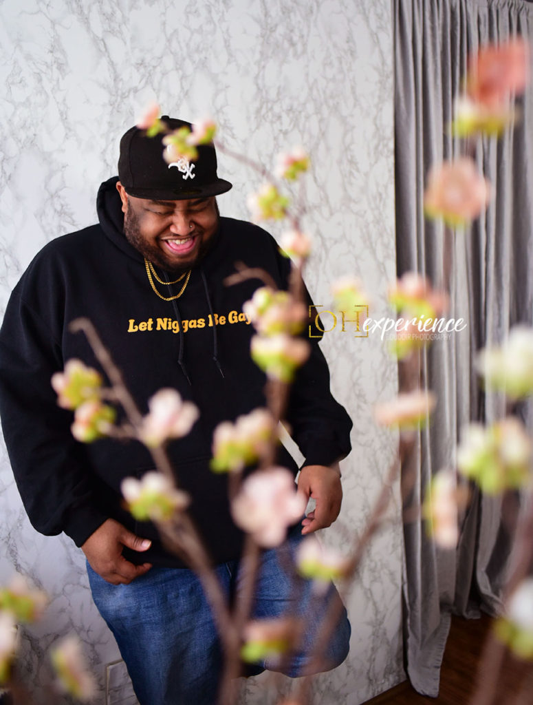 Black man on a marble wall laughing.  His hoodie reads Let Niggas Be Gay. The Negus Project 