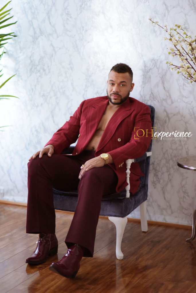 Black man sitting in a chair with a burgundy suit jacket and Burgundy pants. the negus project 