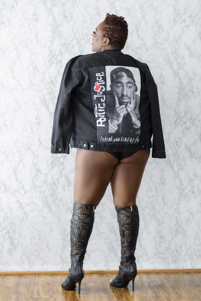 black boudoir photographer, BLACK WOMAN STANDING AGAINST A MARBLE WALL WITH A DENIM JACKET WITH TUPAC ON IT. sHE HAS ON KNEE HIGH BOOTS AND HAS A SHORT NATURAL HAIR CUT