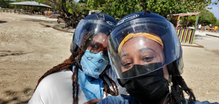 Two black women with helmets on posing for a selfie. One has on a sky blue bandana the other a black face mask