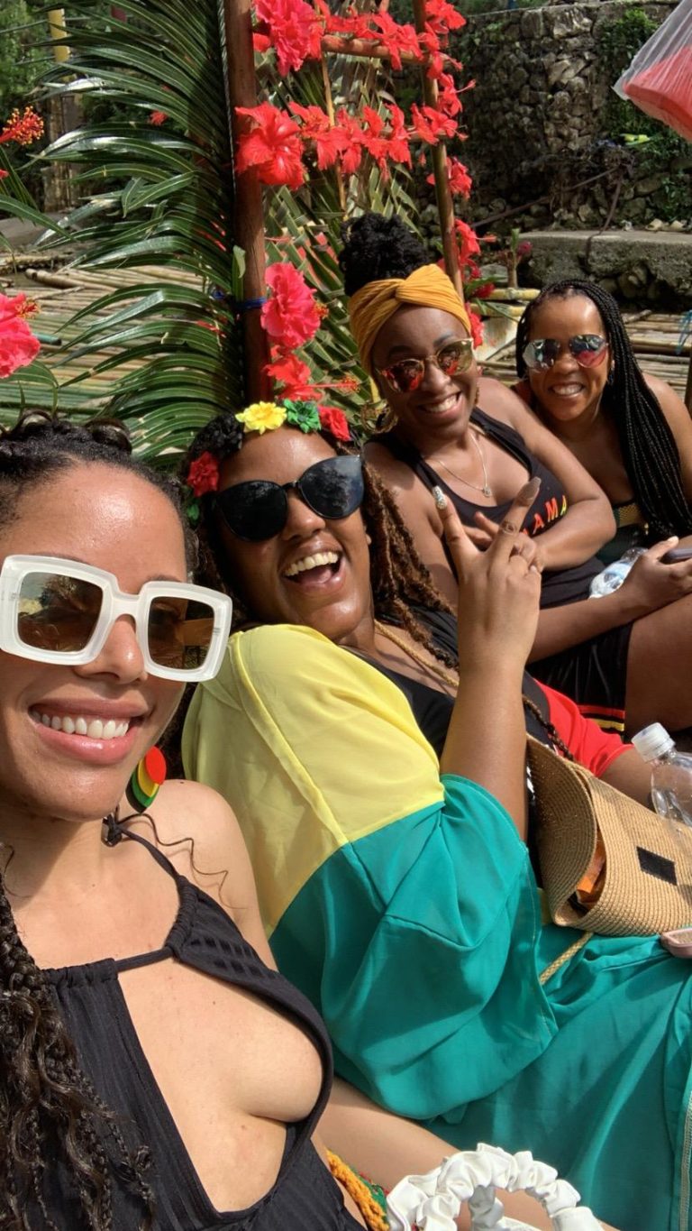 4 black women on a raft in Jamaica, wearing yellow, black and red outfits. All have sunglasses on and one person is holding up the peace sign