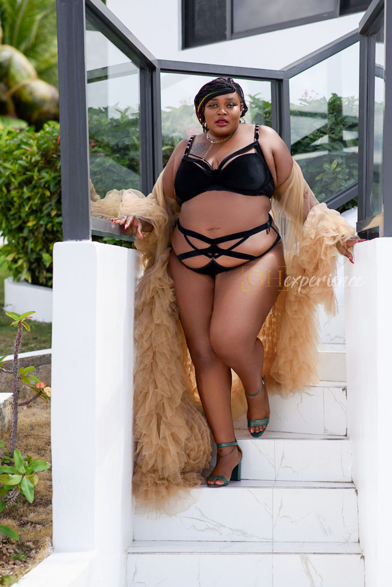PLUS size black woman with box braids in a black velvet bra and panty set. She has on green heels and a luxe tulle sand color robe. She is walking down white steps