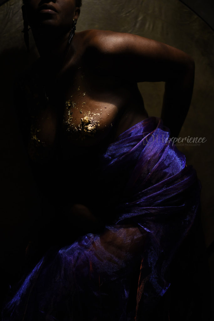 Black woman with gold leaf on her body and large earrings. She has on purple iridescent fabric at her waist