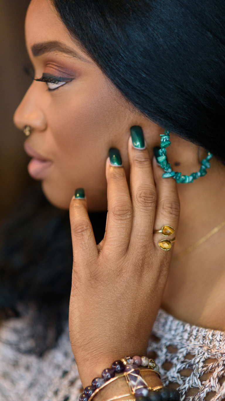 Johari & Lou Black woman with her hand on the side of her face. She has green earrings and bracelets made of gem stones