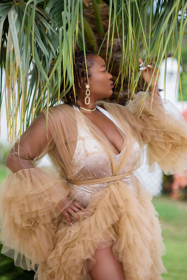 Black woman in a tan color large tulle robe with a velvet halter top body suit. She is standing in front of a palm tree on montego bay jamaica
