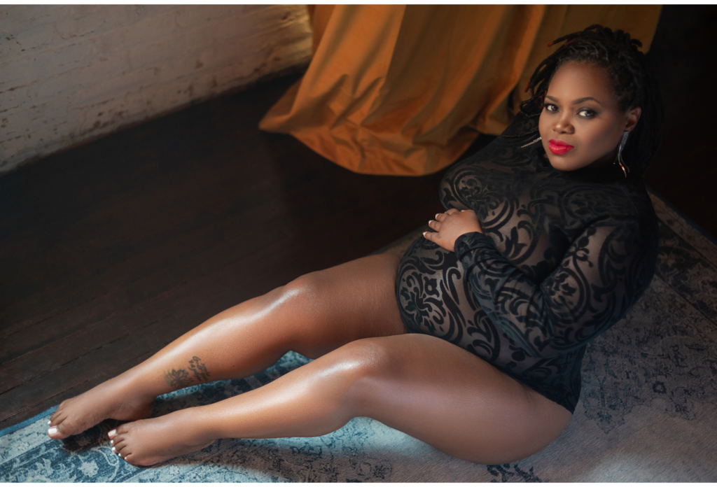 Black pregnant woman dressed in a lace black body suit posed on the floor. Her hand is on her pregnant belly