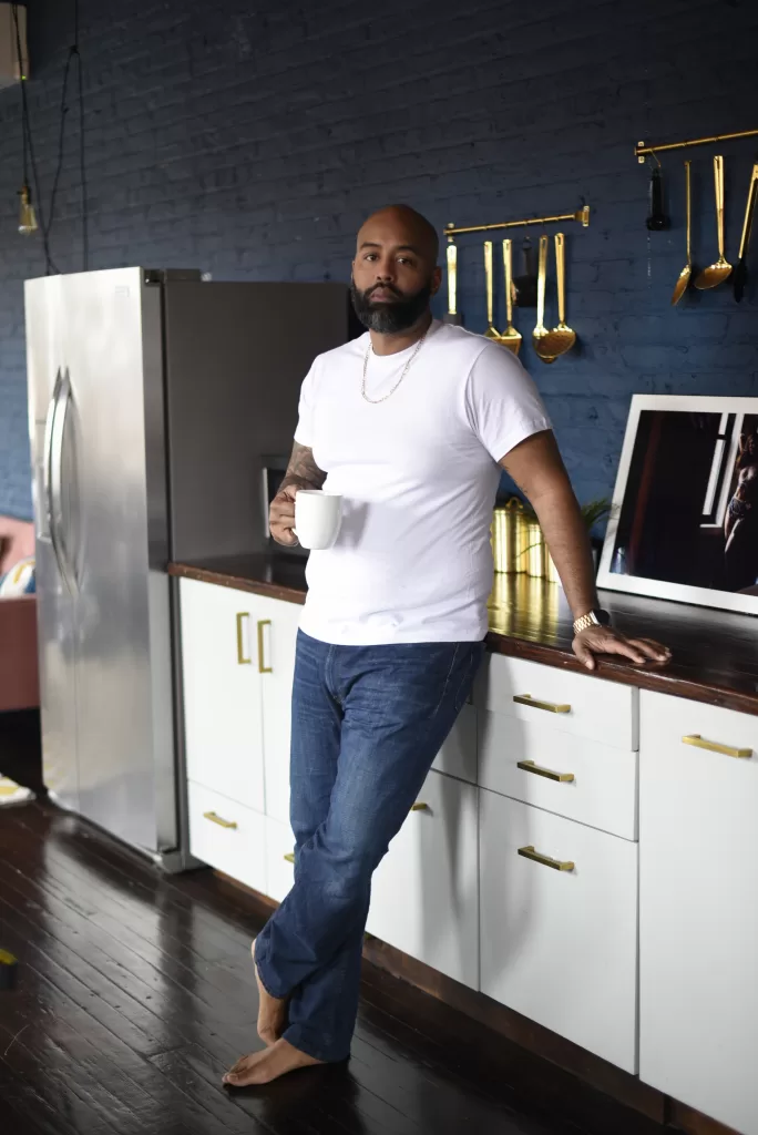 Black man in white shirt with jeans standing with bare feet. He is holding a white mug