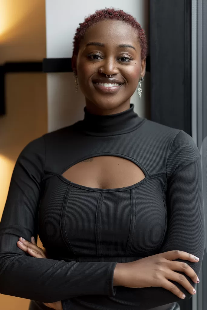 Black woman with a black body suit on. her hands are crossed under her breast and she is smiling.