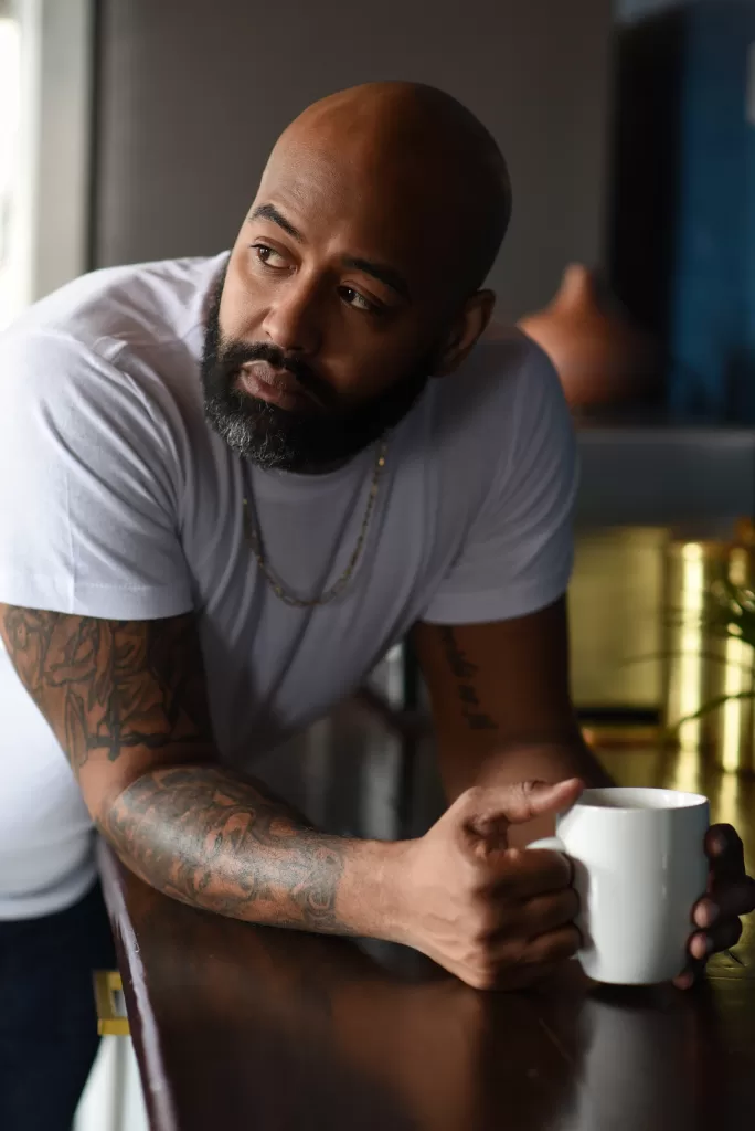 Black man in white t shirt with tattoos on his arm. He has a white mug in his hand