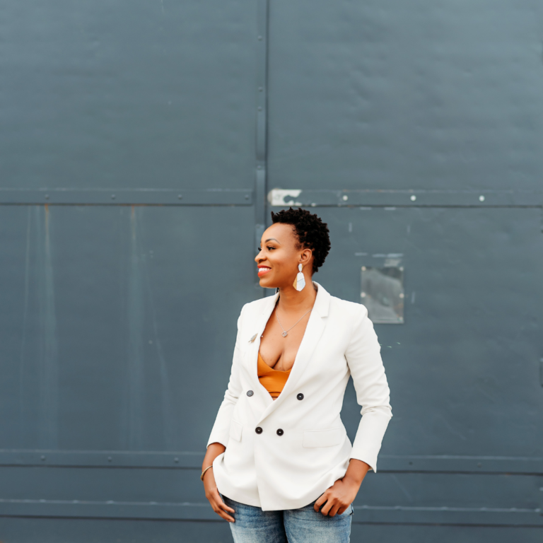 Black woman in white jacket and jeans standing in front of metal large metal door