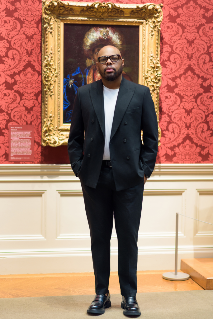Black man in a suit standing in front of a large painting with a gold ornate frame